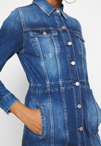 Denimklänning i medelblått, med krage, knappstängning fram, bröstfickor och elastisk midja för en figurnära silhuett.