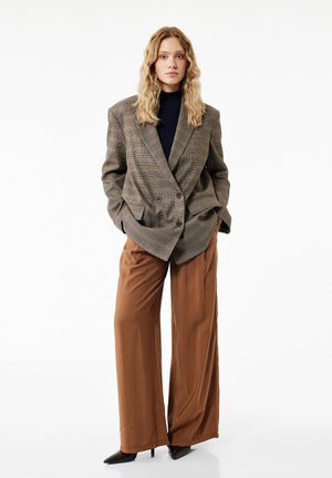 Kvinde med langt bølget hår iført en ternet oversized blazer, sort top, brede brune bukser og sorte spidse hæle, stående mod hvid baggrund.