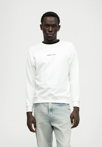 Baltā krekliņa ar apaļu kakla izgriezumu un ribotām piedurknēm, ar nelielu melnu "REPLAY" logo priekšā. Apvienots ar gaiši ziliem džinsiem.