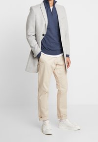 Manteau gris clair sur un pull à fermeture éclair bleu foncé, t-shirt blanc et chinos beige. Des baskets blanches complètent l'ensemble. Design ajusté.