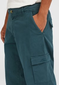 Teal cargo bukser med en glat tekstur, der har sidelommer og et mærketikette. Knaplukning i taljen.
