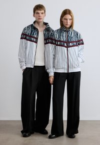 Λευκά μπουφάν με φερμουάρ, διακοσμημένα με μπλε και πράσινα σχέδια και μπορντό ρίγες, συνδυασμένα με φαρδιά μαύρα παντελόνια και μαύρα παπούτσια.