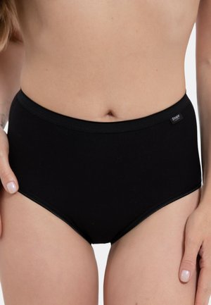 Frau trägt schwarze, hoch taillierte Slip-Unterwäsche mit kleinem "Shape" Logo-Etikett am Bund vor einfachem Hintergrund.