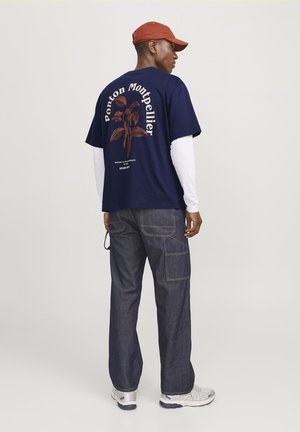 T-shirt in cotone blu navy con logo frontale e grafica di piante, sovrapposto a una maglietta bianca a maniche lunghe; abbinato a pantaloni cargo in denim scuri.
