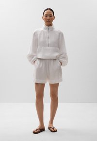 Leinenpullover mit hohem Kragen und halbem Reißverschluss, kombiniert mit passenden Leinen-Shorts. Beide in Weiß, mit lockerer Passform und strukturiertem Stoff.