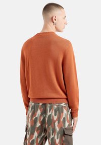 Oranger Strickpullover mit einem gerippten Rundhalsausschnitt und Saum. Kombiniert mit camouflagefarbigen Cargo-Hosen, die Taschen und eine Mischung aus Erdtonen aufweisen.
