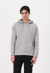 Haut à capuche gris avec une poche avant, un cordon de serrage à la capuche et des poignets côtelés. Présente une texture douce et un design simple. Porté avec un pantalon noir.