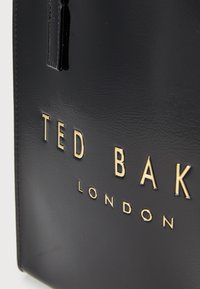 Sac en cuir noir avec un logo embosser en lettres dorées, finition texturée et forme rectangulaire élégante.