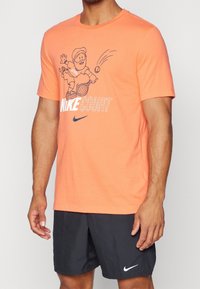 Mies, joka pukeutuu oranssiin Nike-t-paitaan, jossa on sarjakuvamainen tennispelaaja graafinen kuva, sekä mustiin Nike-shortseihin, joissa on valkoinen swoosh-logo.