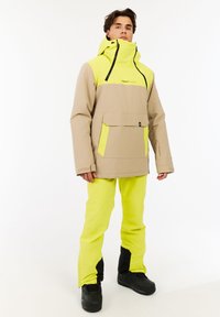 Heren skijack in beige en neon geel. Kenmerken zijn een voorvak, verstelbare capuchon, zwarte ritsen en bijpassende neon broek met zwarte accenten.