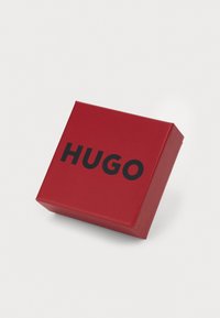 Boîte rectangulaire rouge avec une finition brillante, portant le mot "HUGO" en lettres noires épaisses à l'avant.