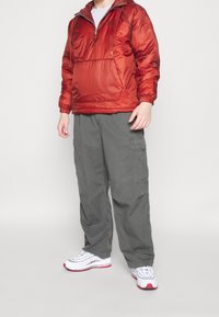 Röd, lätt, huvad midja med halvdragkedja och elastiska ärmmuddar, i kombination med grå cargobyxor och vita sneakers med röda detaljer.