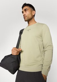 Lys grøn sweatshirt med ribstrikkede manchetter og kant. Har en lille sort "HYROX" logo. Modellen holder en sort træningstaske over skulderen.