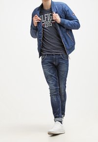 Chaqueta bomber azul marino con cuello acanalado, camiseta gráfica gris, jeans ajustados de un azul medio y zapatillas altas blancas.