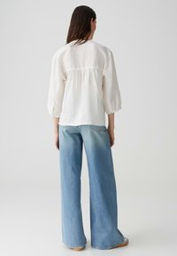 Blusa bianca in lino con maniche a sbuffo, dettaglio arricciato sul retro, abbinata a jeans a gamba larga di colore azzurro chiaro, con lavaggio chiaro e tasche frontali.