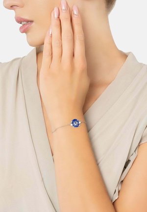 Silver bracelet featuring a round blue enamel charm with an eye design, set against a skin tone, worn on a bare wrist.