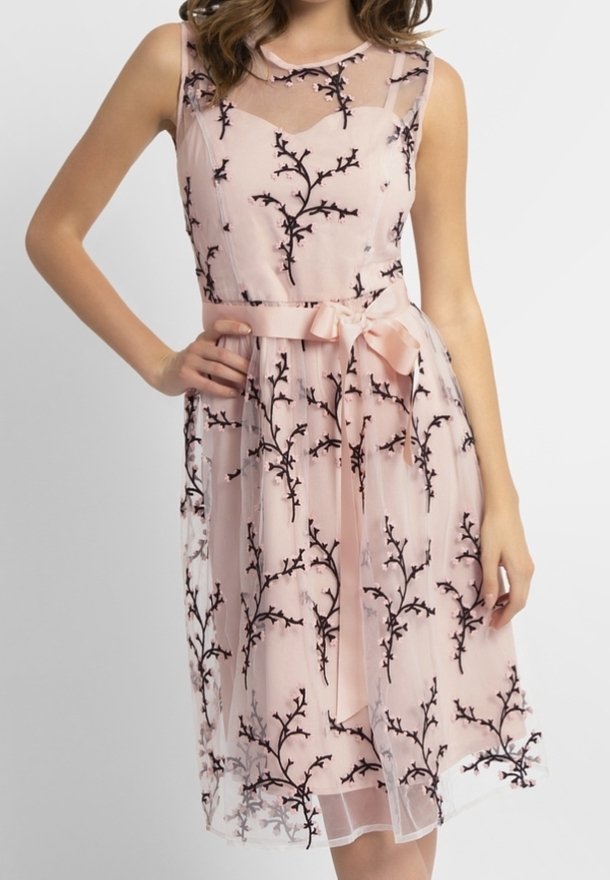 Robe rose sans manches avec broderie florale noire, encolure transparente et ruban noué à la taille, portée par une femme la main sur la hanche.
