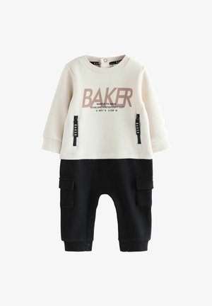 Kaksiosainen asu kerma- ja mustassa, jossa on suuri "BAKER"-logo, vetoketjulliset taskut ja nappisuljennat nilkoissa. Pehmeä kangas.