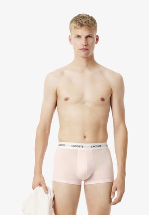 Pink cotton boxer briefs with a white waistband featuring the Lacoste logo, worn by a shirtless male model holding a light-colored shirt.