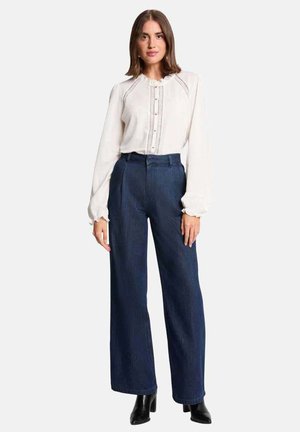 Femme portant un chemisier blanc à manches longues avec des détails en dentelle, un jean large taille haute foncé et des bottines noires, debout devant un fond blanc.