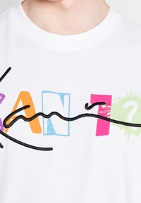 Camiseta de algodón blanca con gráfico de texto multicolor "KAN" acompañado de formas juguetonas, acentuado con líneas bordadas en negro y diseños de salpicaduras.