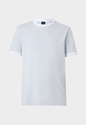 Kvinde og mand står op ad en hvid væg, begge iført lyseblå T-shirts og sorte underdele; kvinden holder en sort håndtaske, manden har et armbånd.