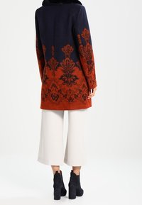 Lang uldfrakke med en marineblå overdel og en orange underdel, prydet med sorte blomsterdesigns. Sat sammen med cremefarvede brede bukser og sorte ankelstøvler.