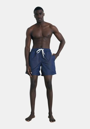 Costume da bagno blu navy con cintura regolabile, caratterizzati da una texture liscia e una vestibilità comoda, indossati da un modello maschile in piedi a piedi nudi.