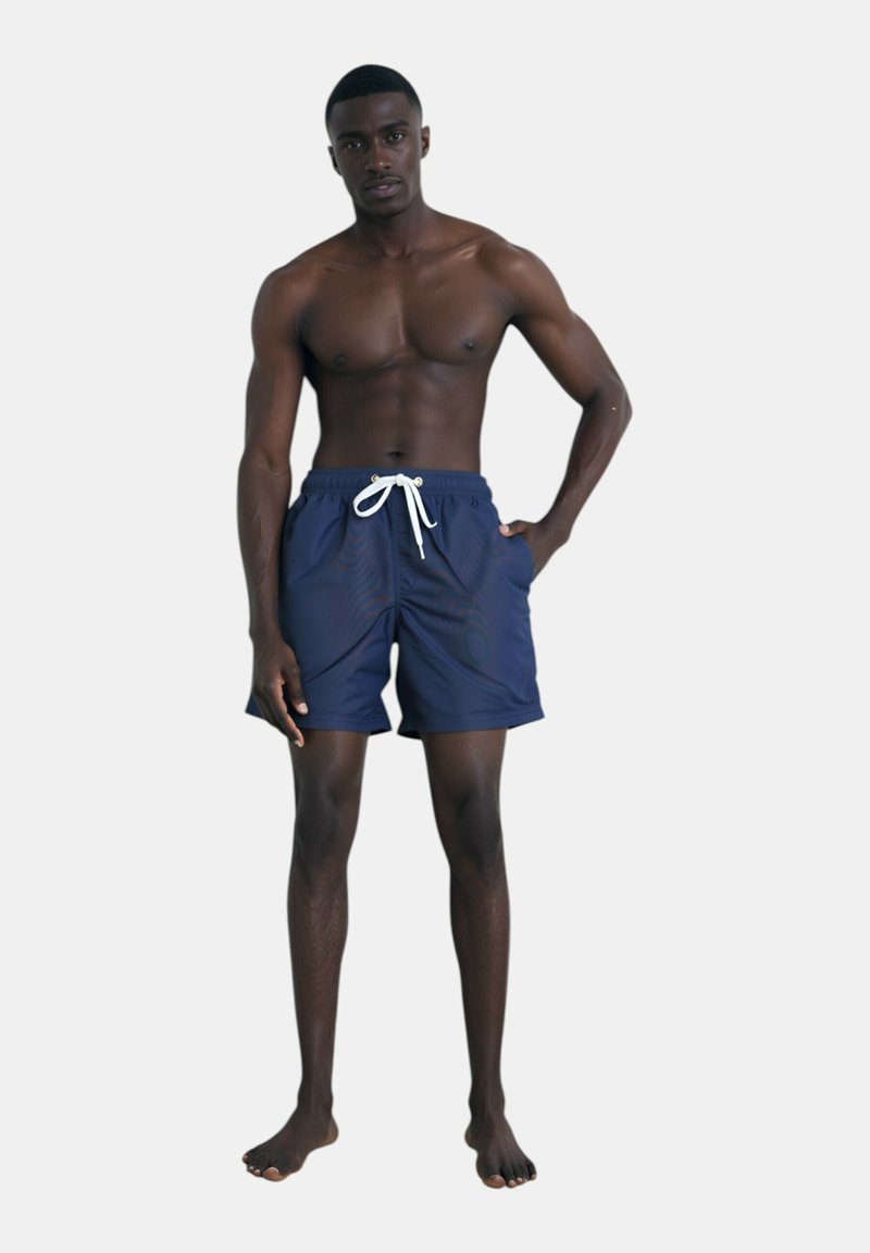 Costume da bagno blu navy con cintura regolabile, caratterizzati da una texture liscia e una vestibilità comoda, indossati da un modello maschile in piedi a piedi nudi.