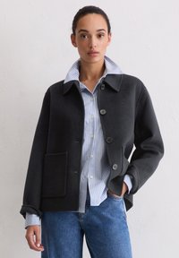 Czarna wełniana kurtka z klasycznym kołnierzem, zapinana na guziki z przodu, z jedną kieszenią na piersi, noszona na niebieskiej koszuli w paski i dżinsach.