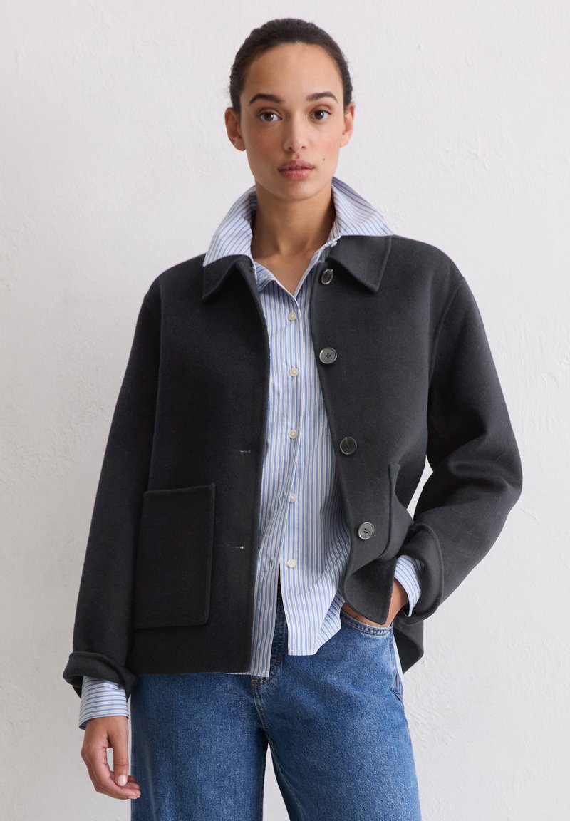 Czarna wełniana kurtka z klasycznym kołnierzem, zapinana na guziki z przodu, z jedną kieszenią na piersi, noszona na niebieskiej koszuli w paski i dżinsach.