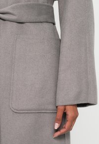 Harmaa villatakki, jossa on suuri sivutasku, leveät hihat ja pehmeän tuntuinen vyötärönauha samasta materiaalista. Saumalistaus näkyvissä.