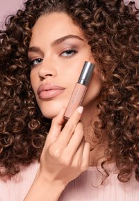 Płynny produkt do ust w szklanym opakowaniu z srebrną nakrętką; delikatny różowy odcień; modelka trzyma go przy twarzy, prezentując błyszczące usta.