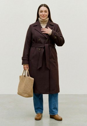 Une femme porte un manteau marron foncé ceinturé sur un col roulé beige et un jean, et tient un sac cabas tissé devant un fond de studio uni.