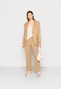 Blazer bege ajustado e calças combinando em um tecido suave. O modelo usa uma blusa creme e uma bolsa branca com detalhes em corrente, combinadas com sandálias brancas.