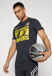 Camiseta de algodón negra con gráfico amarillo de "Los Angeles Lakers". Mangas cortas, cuello redondo, combinada con pantalones cortos de malla negros, sosteniendo un balón de baloncesto.