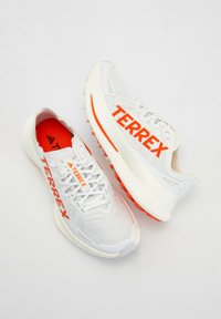 Paire de chaussures de course Terrex blanches avec semelles orange et marquage orange vif sur les côtés et les languettes, sur fond blanc.
