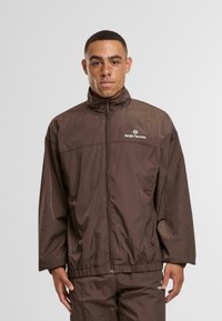 Moški s kratkimi lasmi in pegami, oblečen v rjavo jakno na zadrgo Sergio Tacchini in ujemajoče se hlače, proti enotni ozadju.