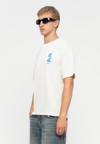 White cotton t-shirt with a relaxed fit, featuring a blue graphic design on the left side. Model wears black sunglasses. Jeans are faded blue.