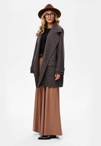 Bruine houndstooth jas met grote revers, zijzakken; gedragen over een zwarte top en beige wijde broek, gecombineerd met een bruine hoed en zwarte laarzen.