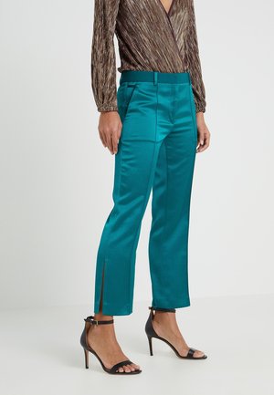 Türkisfarbene Satin-Cropped-Hosen mit vorderen Bügelfalten und Knöchelschlitzen, kombiniert mit schwarzen High-Heel-Sandalen mit Knöchelriemen und einem metallischen Langarmtop.