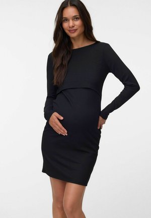 Femme enceinte aux longs cheveux foncés porte une robe noire ajustée à manches longues, tenant doucement son ventre, debout devant un fond blanc uni.