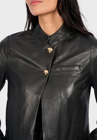 Black leather jacket featuring a high collar, two gold buttons, and a small chest pocket with a smooth, shiny texture.