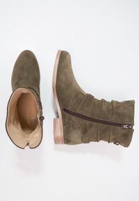 Bottine en daim vert olive avec un bout arrondi, un design froissé, une fermeture à glissière latérale et un talon en bois. Intérieur doublé de tissu beige.