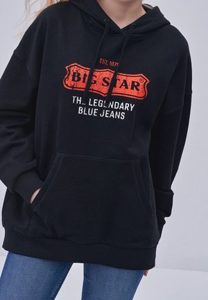 Osoba nosząca czarną bluzę z kapturem z logo „BIESTAR The Legendary Blue Jeans” w czerwieni i bieli, w zestawieniu z niebieskimi dżinsami.