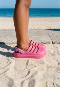 Ben iført lyserøde slip-on sandaler med lavendelrem, stående på sand nær havet og en betonsti på en solrig dag.