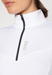 Bjela zip-up pulover s visokim ovratnikom, s crnim patentnim zatvaračem i sivo "COLMAR" logotipom. Glatka tkanina s blago prilagođenim dizajnom.