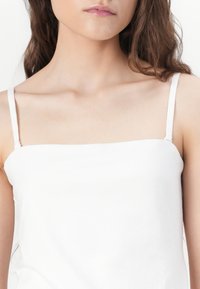Biely top s tenkými nastaviteľnými ramienkami, s hladkou textúrou a rovnými okrajmi výstrihu, zameraný na minimalistické dizajnové detaily.