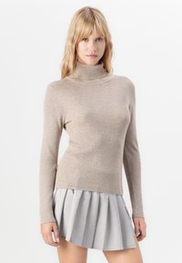 Kvinde med langt blondt hår iført en beigefarvet ribbestrikket turtleneck sweater og en lys grå plisseret miniskirt mod en hvid baggrund.