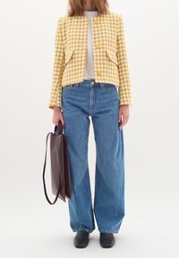 Gul och vit rutmönstrad jacka med två framfickor, i kombination med blå högmidjade jeans och svarta skor, hållande en brun handväska.
